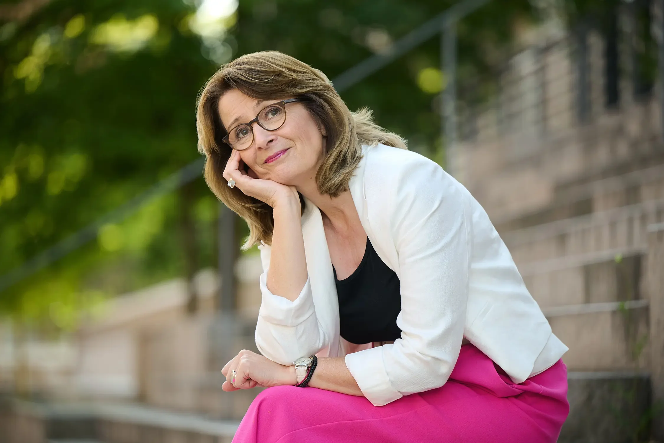 Brigitta Wurnig mit Brille sitzt auf einer Parkbank, trägt einen weißen Blazer, schwarze Bluse und pinke Hose, und schaut nachdenklich in die Kamera, umgeben von grünen Bäumen.