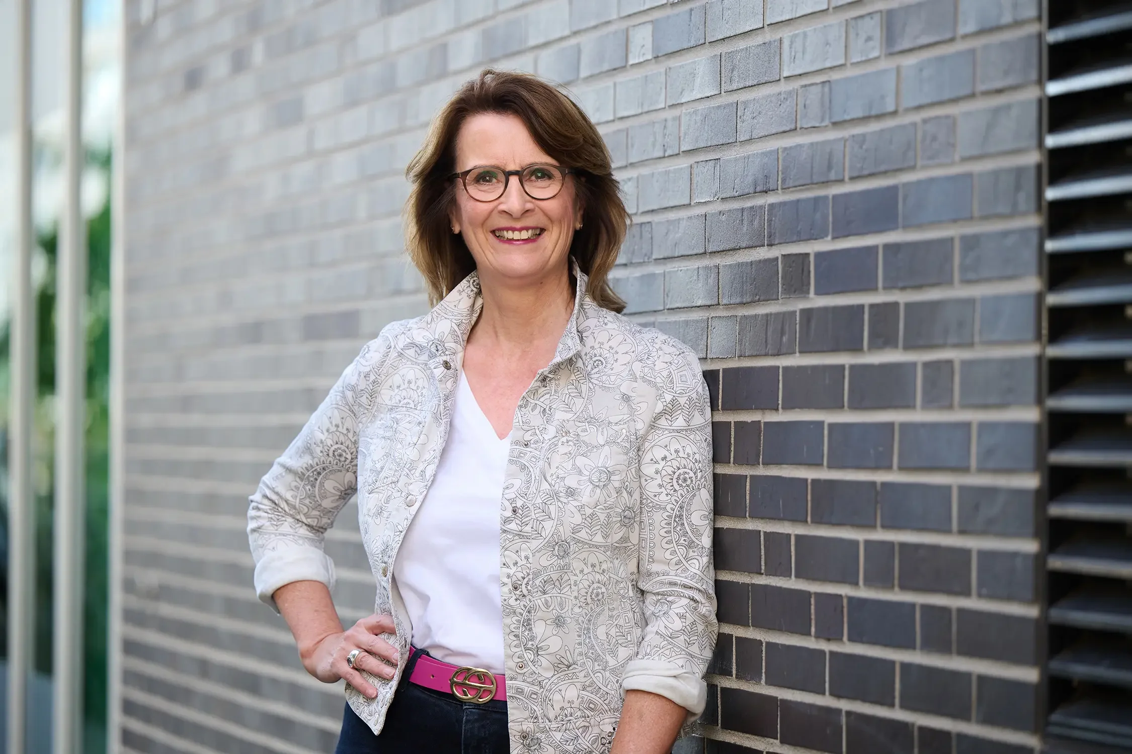 Eine lächelnde Brigitta Wurnig mit Brille und braunen Haaren lehnt an einer gemauerten Wand im Freien, trägt eine weiße T-Shirt und eine Jacke mit floralem Muster, dazu einen pinken Gürtel.