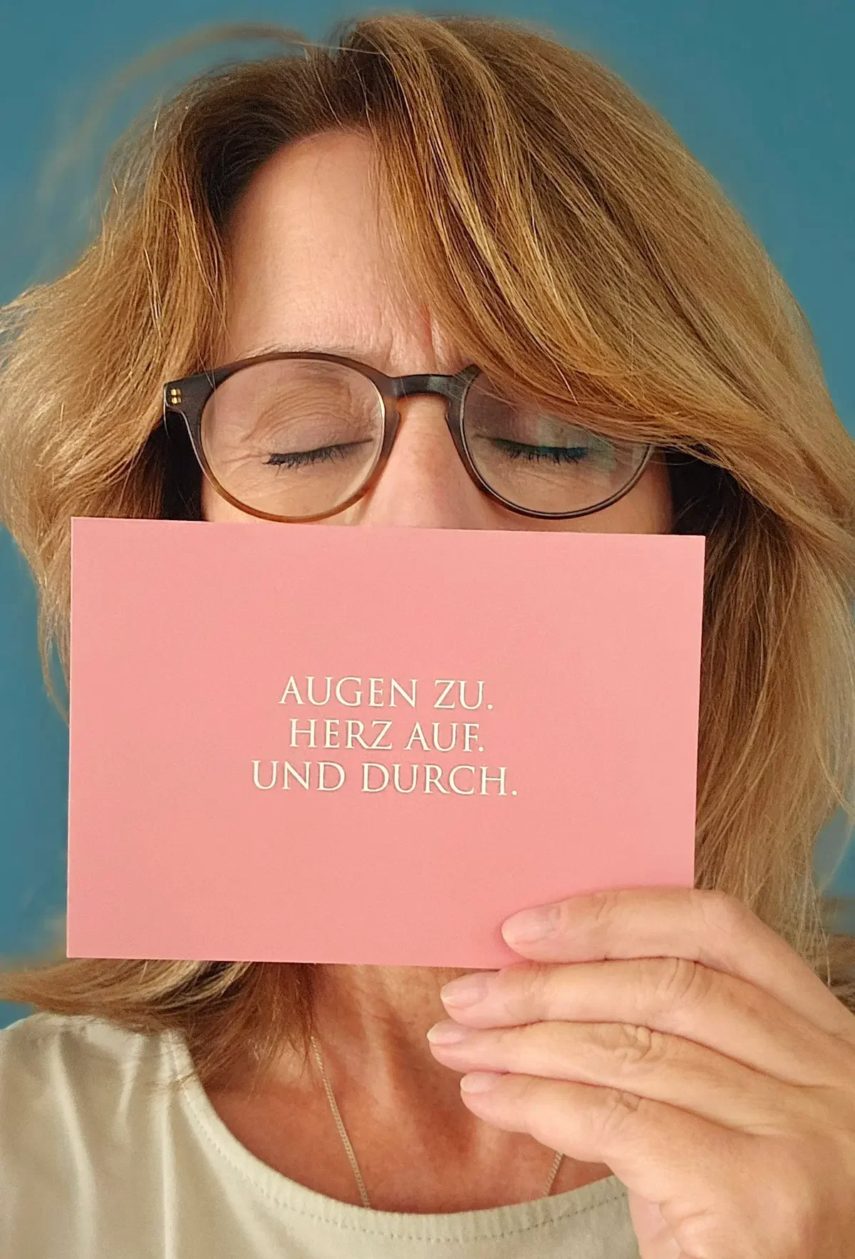 Brigitta Wurnig mit lockigem braunem Haar, die eine Brille trägt, hält ein rosa Karte vor ihr Gesicht, auf der steht: "Augen zu. Herz auf. Und durch."