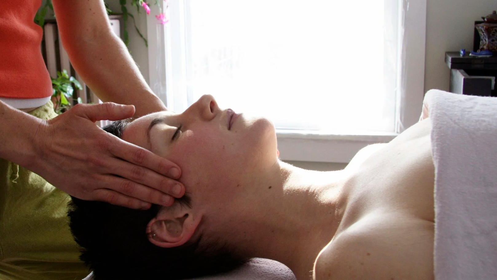 A person receiving a head massage or chiropractor treatment while lying down with eyes closed in a bright room.