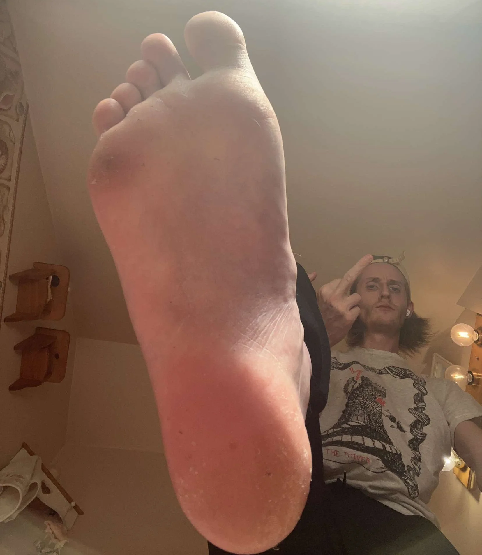 Close-up of a foot with a person giving a middle finger gesture in the background, wearing a graphic T-shirt and cap.