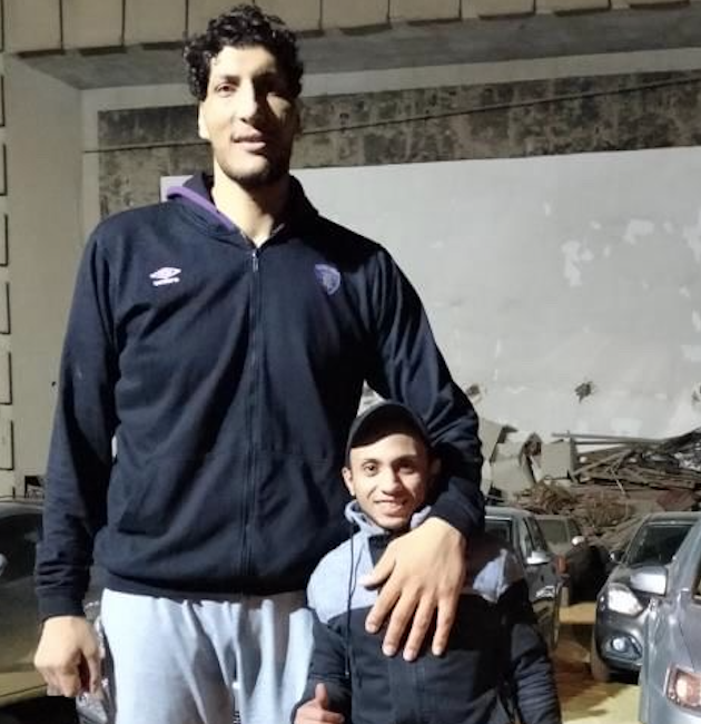 Two men posing for a photo in an outdoor setting, with cars parked in the background. The man on the left is significantly taller, wearing a black jacket and sweatpants. The man on the right is shorter, wearing a cap and black jacket with gray accents.