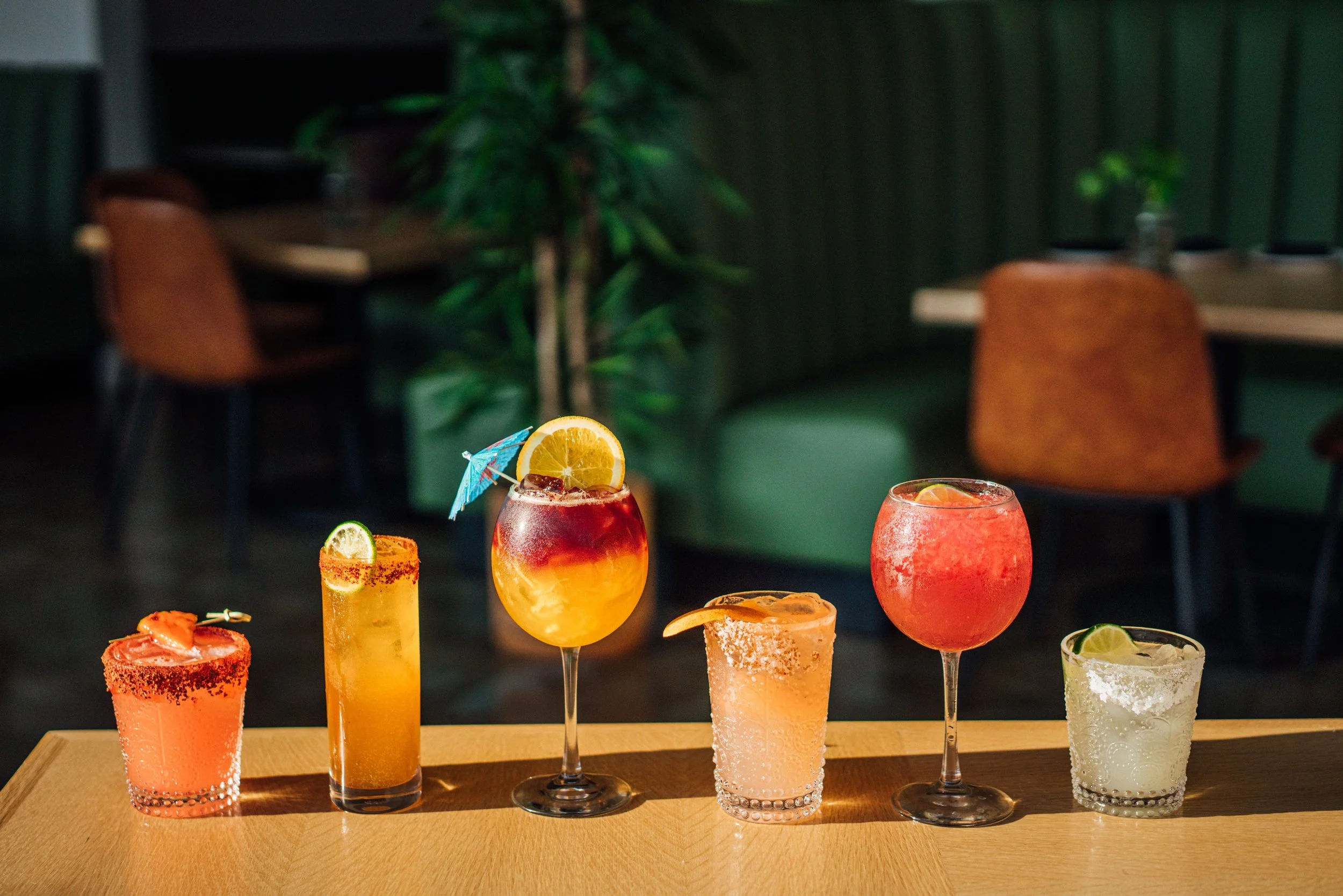Six colorful cocktails on a wooden table in a dimly lit restaurant or bar setting.
