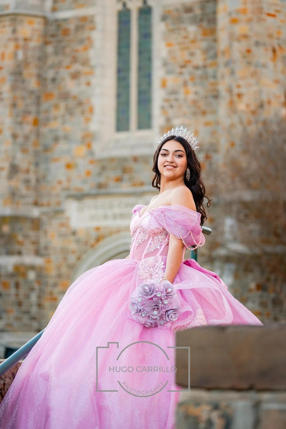 Young woman in a pink ball gown with off-shoulder sleeves, holding a bouquet of pink roses, wearing a tiara and earrings, standing outside a brick church building.