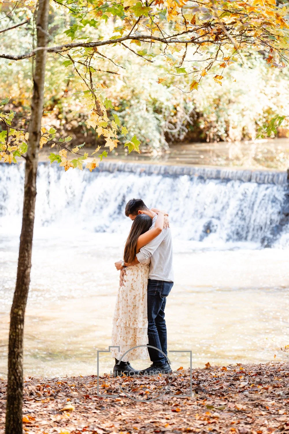 A couple embracing by a waterfall in a forest with autumn leaves.