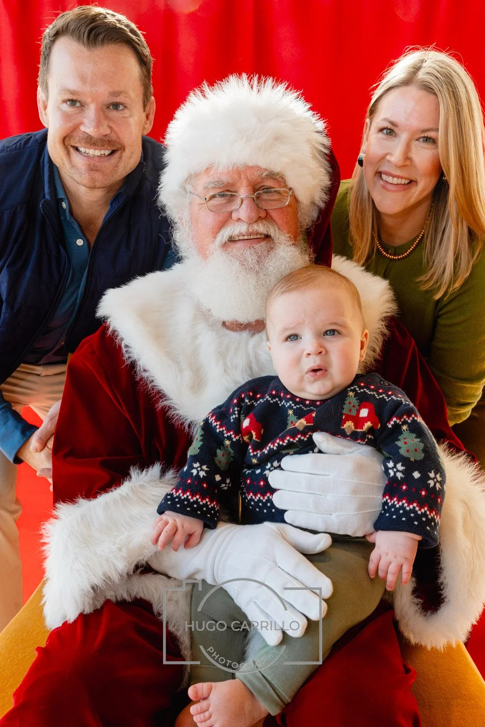 Family photo with Santa Claus, a young child, and two adults, all smiling, against a red backdrop.