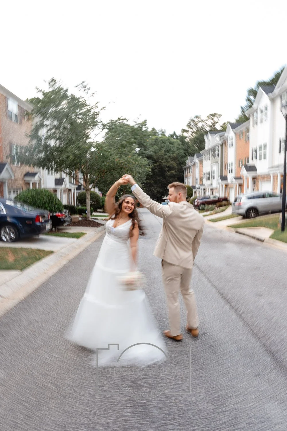 A bride and groom dancing in the middle of a residential street lined with houses and parked cars.