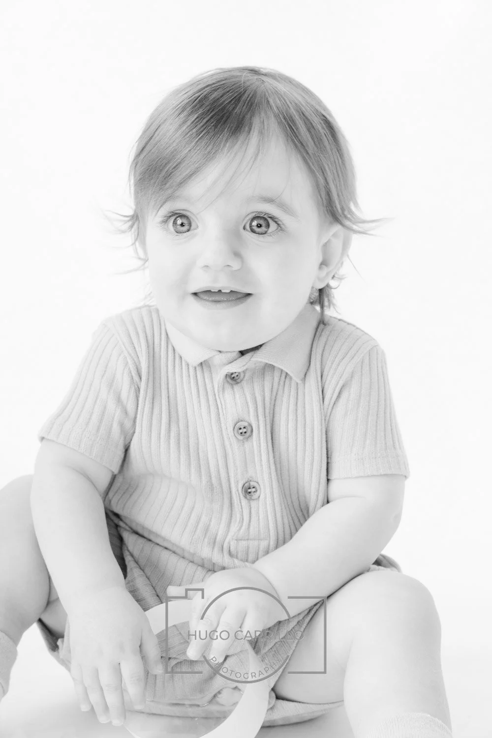 Black and white photo of a young child with light-colored hair, large eyes, smiling, wearing a striped polo shirt and shorts.