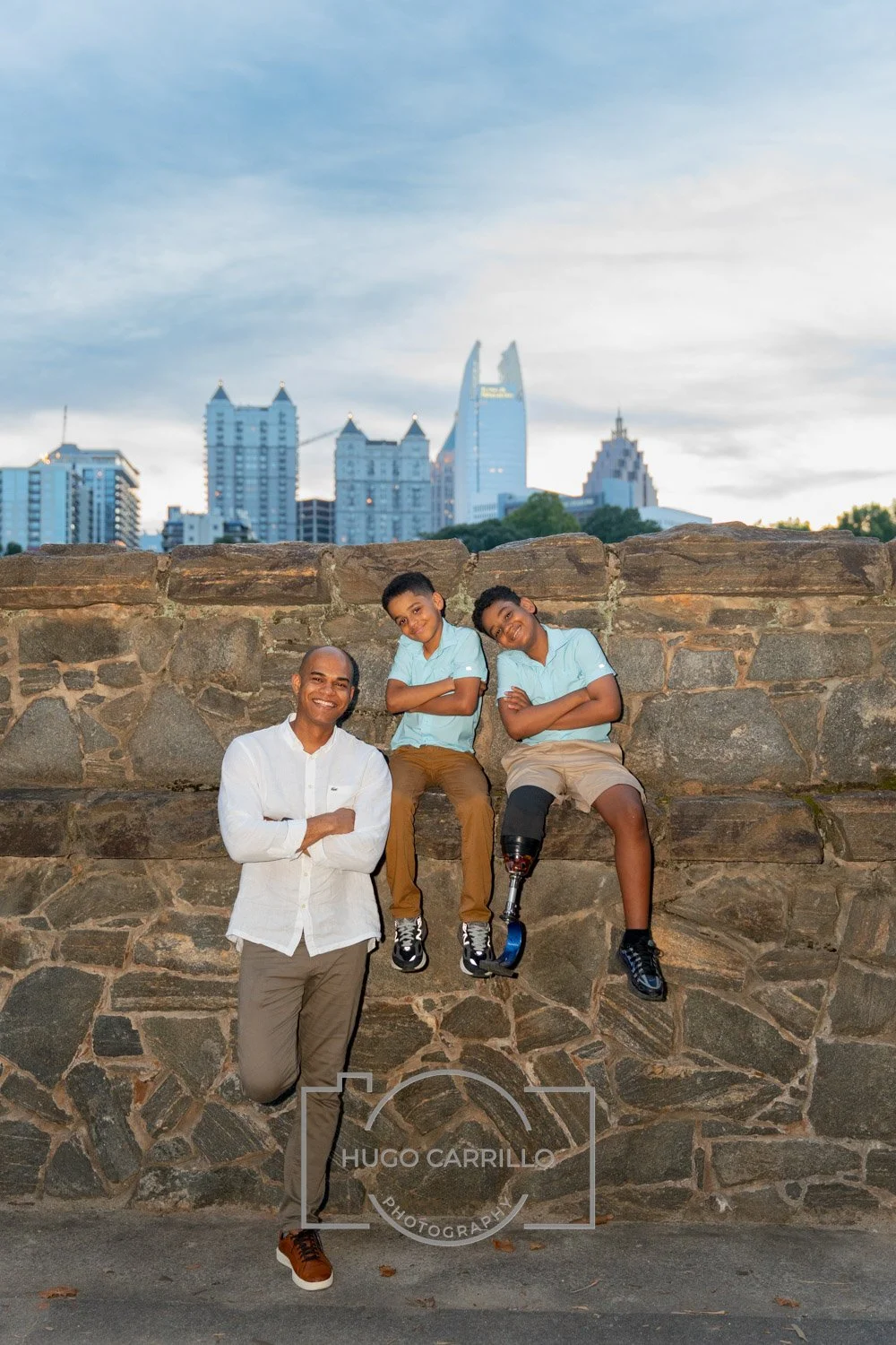 A man with a prosthetic leg and two boys are sitting on a stone wall with a city skyline in the background. The man and boys are smiling and appear happy.