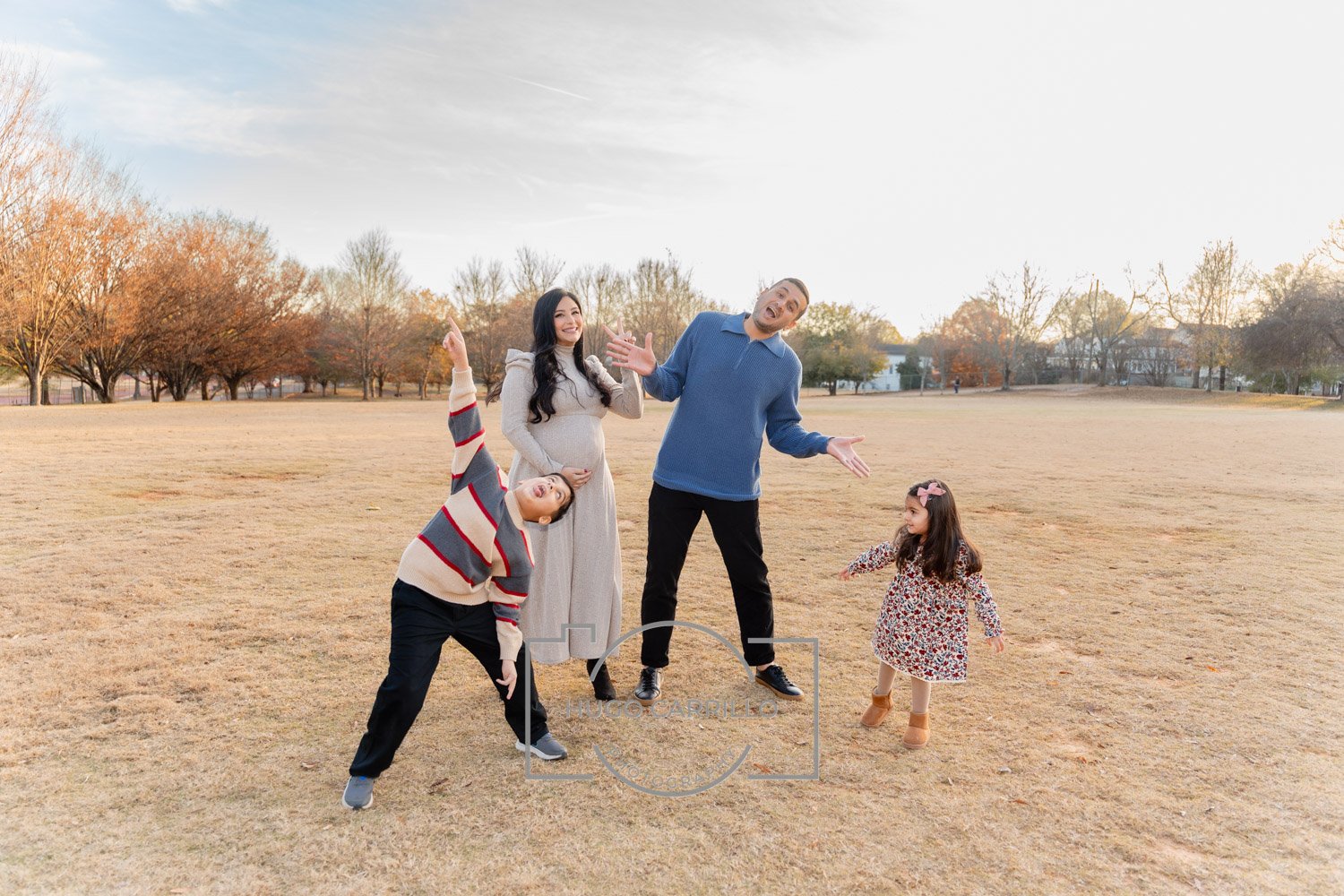 A family of five standing on a grassy field with trees and houses in the background during autumn. The pregnant mother is smiling and wearing a beige dress, the father is gesturing with his hands and wearing a blue sweater, a young girl with a pink b