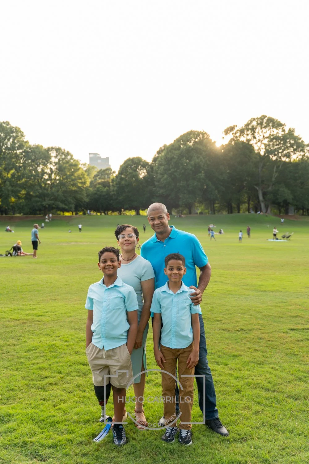 A family of four stands together in a park during golden hour. The adult woman, adult man, and two boys are smiling at the camera, with trees and other park visitors in the background.