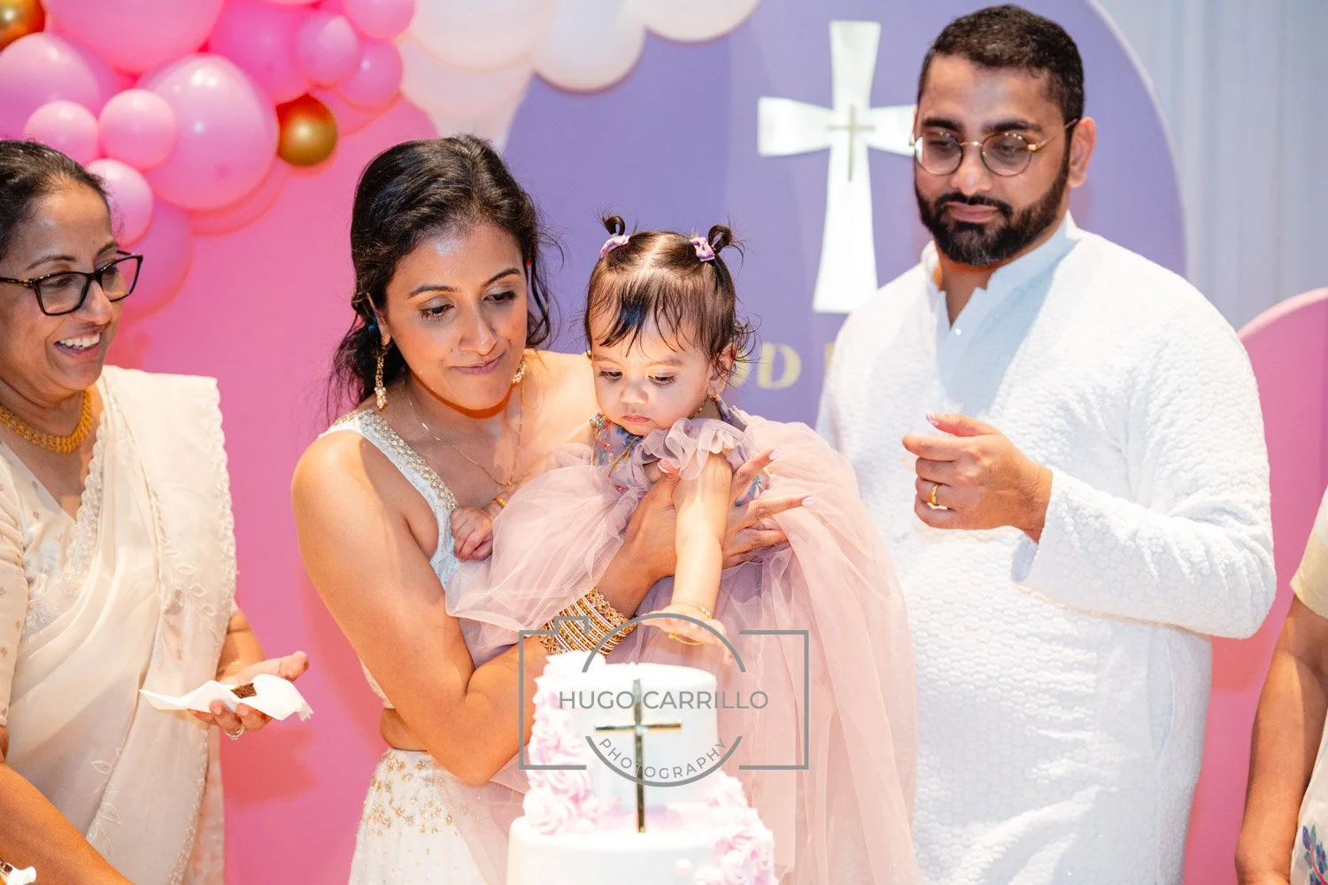 A family at a religious celebration, with a woman holding a young girl dressed in pink, a man in white, and a woman in glasses and traditional attire, with balloon decorations and a cross in the background.