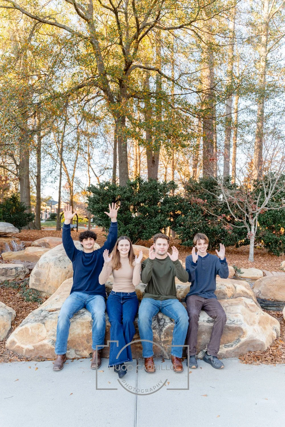 Four young adults sitting on large rocks in a park during autumn, with trees and fallen leaves in the background, waving at the camera.