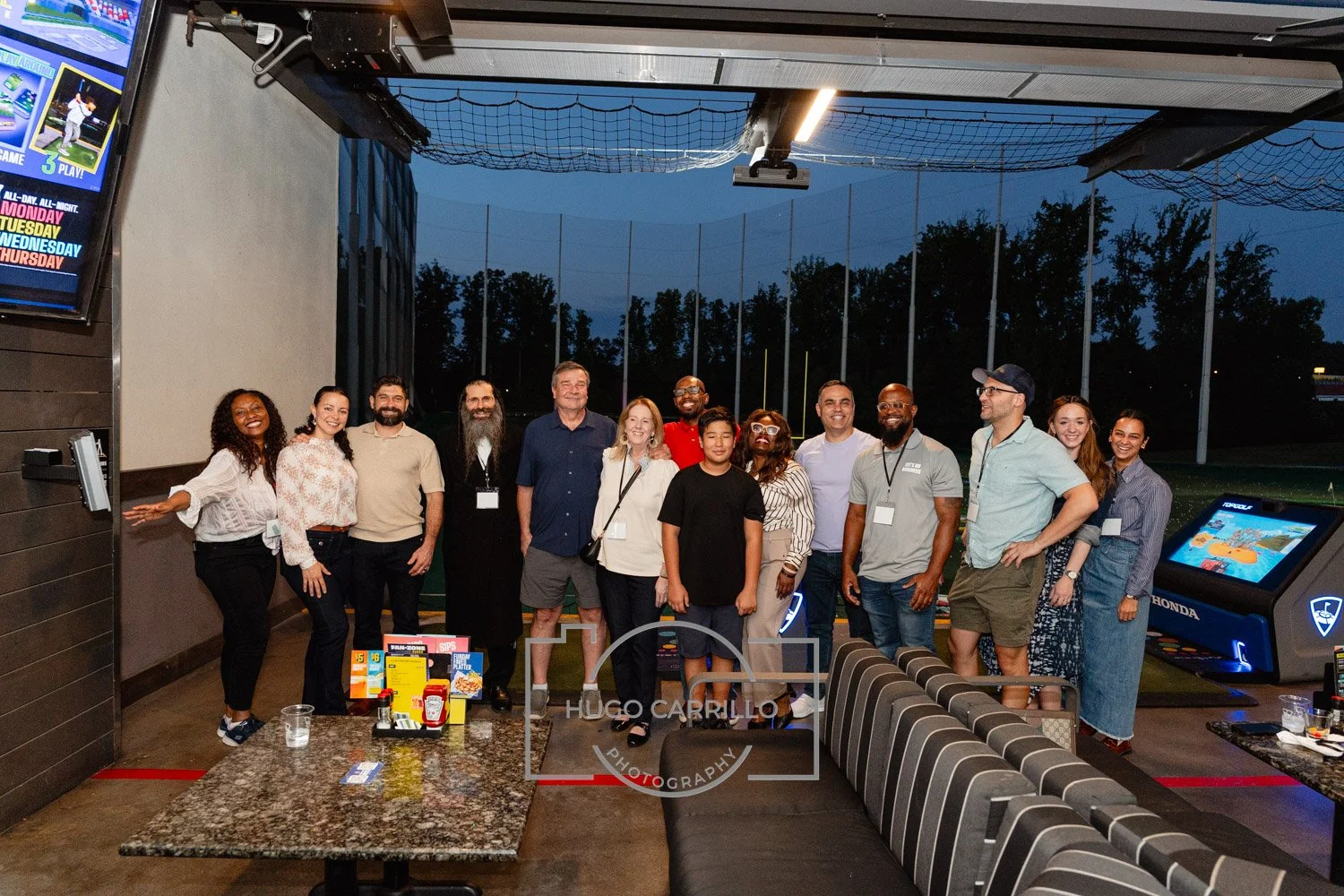 Group of people standing together at a social event in a sports bar or restaurant, with a golf driving range visible in the background during dusk.