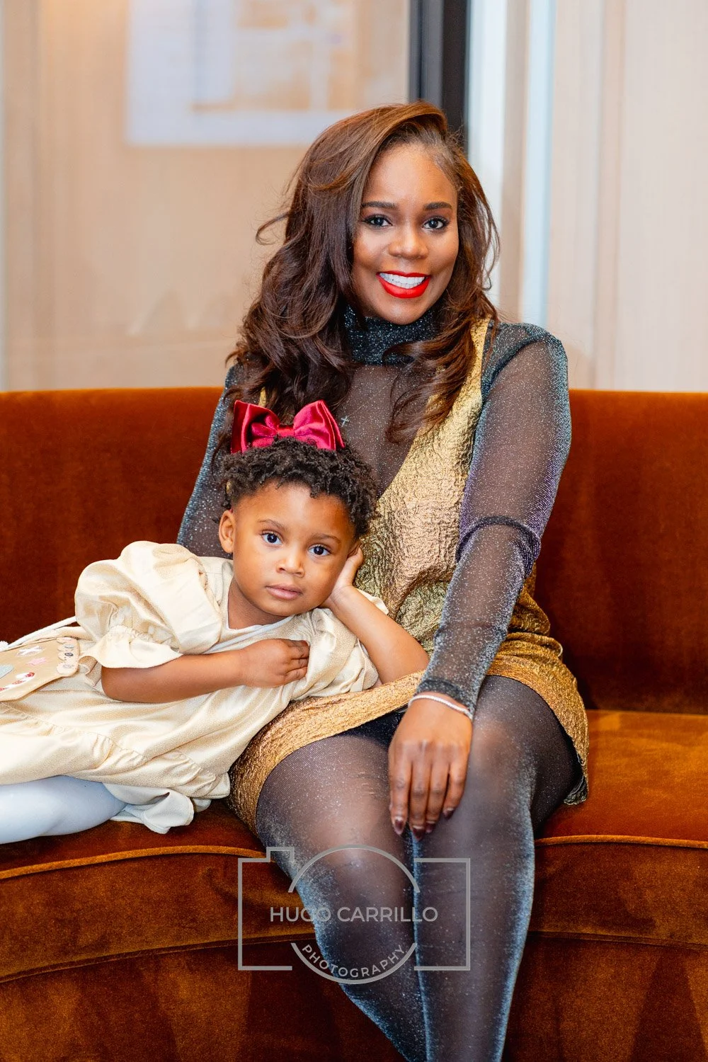 A woman with dark hair wearing a black, gold, and sheer dress sitting on a brown couch, smiling with red lipstick, with a young girl in a cream-colored dress resting her head on her lap and looking at the camera, in an indoor setting.