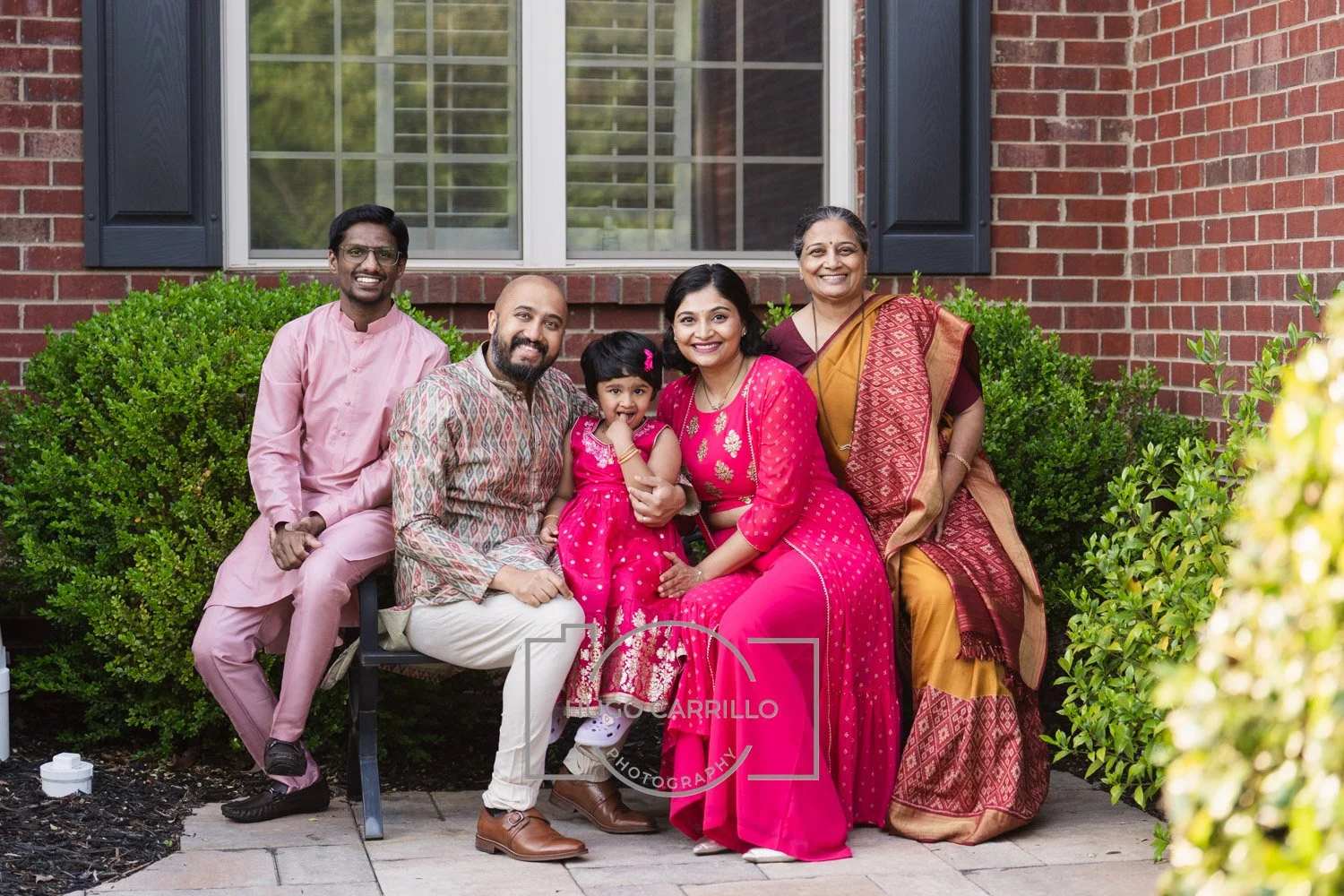 A family of six sitting on a bench outside a brick house, dressed in colorful traditional Indian clothes, smiling and celebrating together.