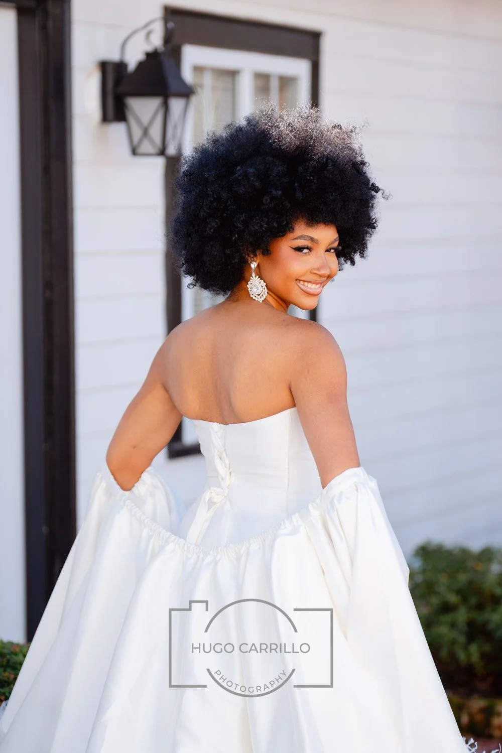 A woman in a white strapless wedding dress, smiling and looking over her shoulder, standing outside near a building with white siding and a black lantern.