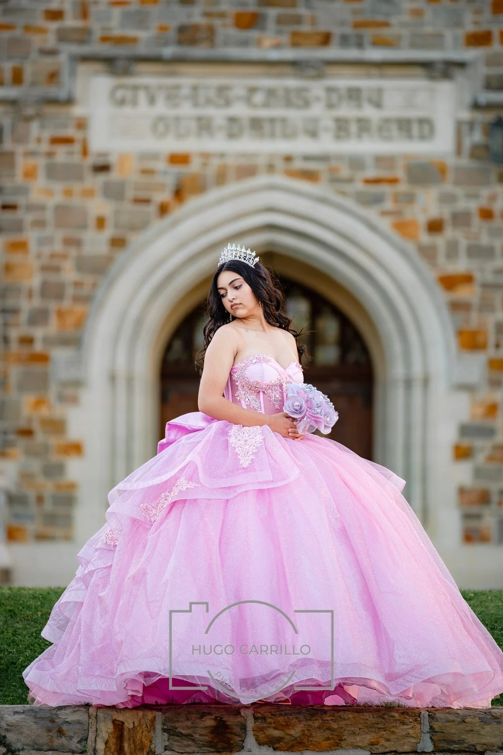 Young woman in a pink quinceañera dress holding a bouquet of flowers, wearing a tiara, standing in front of a brick building with an arched doorway.