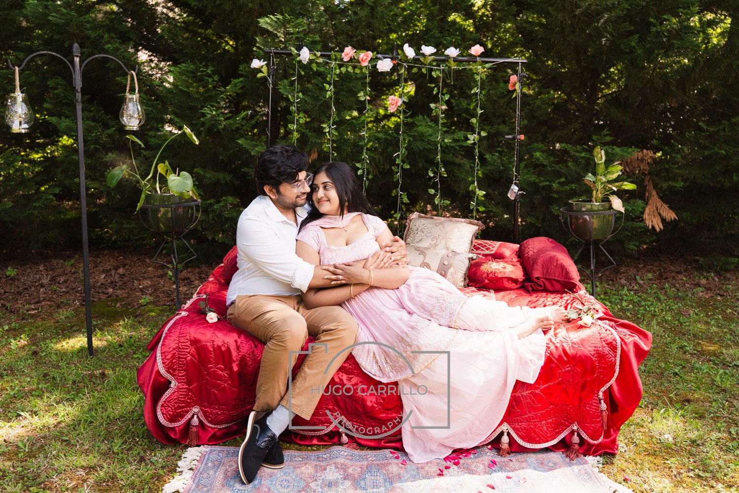 A couple sitting on a red bed in a green outdoor setting, with the man embracing the woman who is lying down, surrounded by pillows and floral decorations.