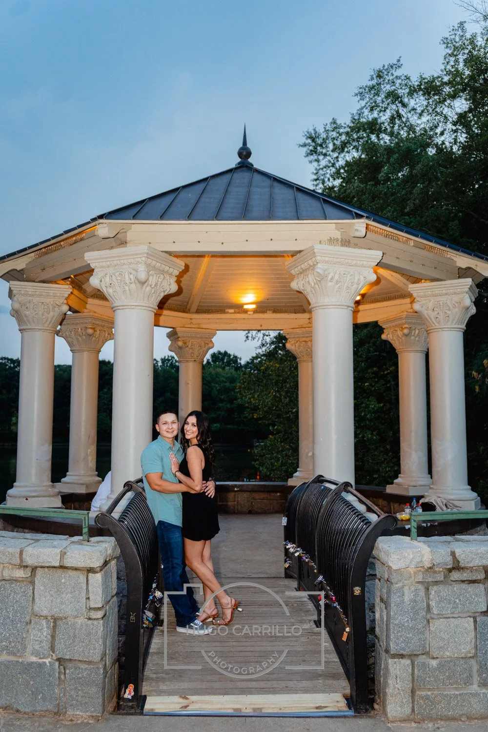 A couple standing and hugging under a classical-style gazebo with white columns, during dusk in a park.