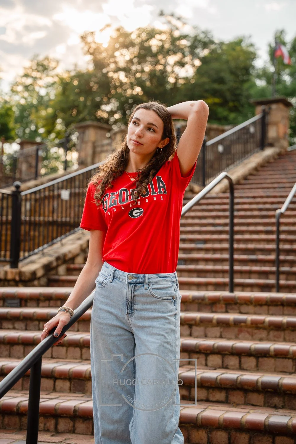 A young woman wearing a red Georgia Bulldogs t-shirt and light blue jeans standing on brick stairs outdoors at sunset, with trees and a flag in the background.