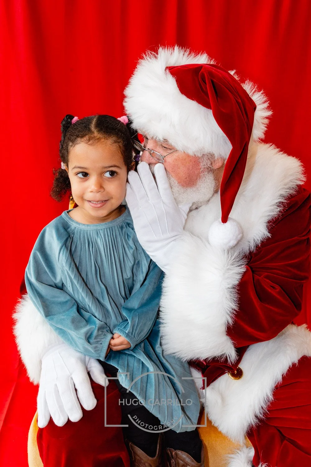 A young girl sits on Santa Claus's lap, listening to him whisper. Santa is in a red velvet suit with white fur trim, and the girl is wearing a blue dress. They are against a red background.