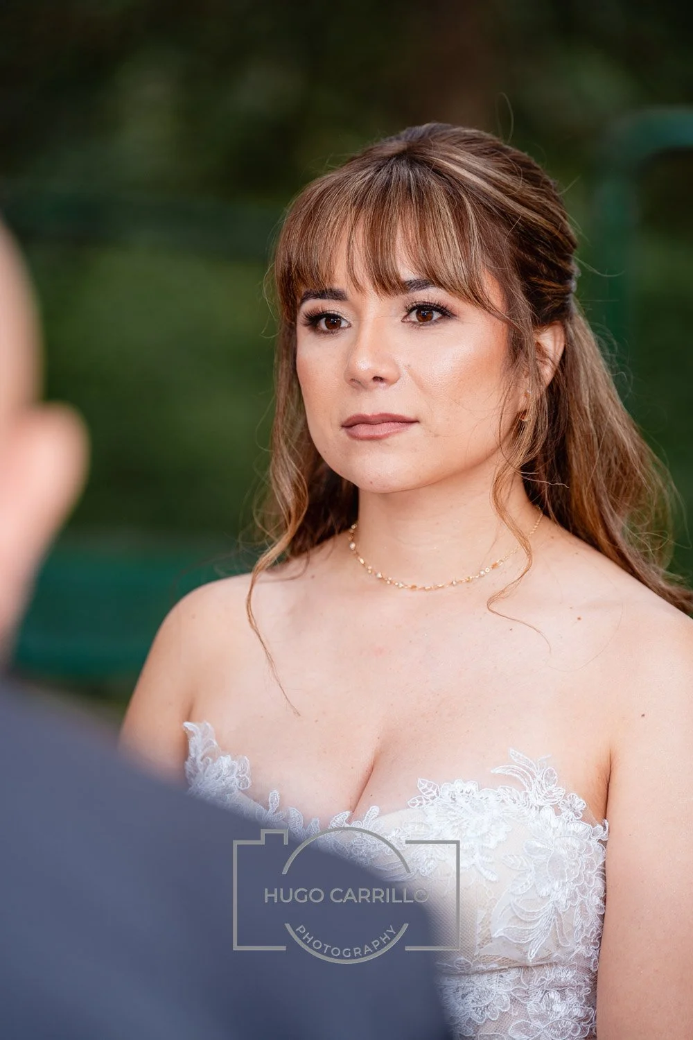 A woman wearing a white lace dress and a delicate necklace, standing outdoors with blurred greenery in the background.