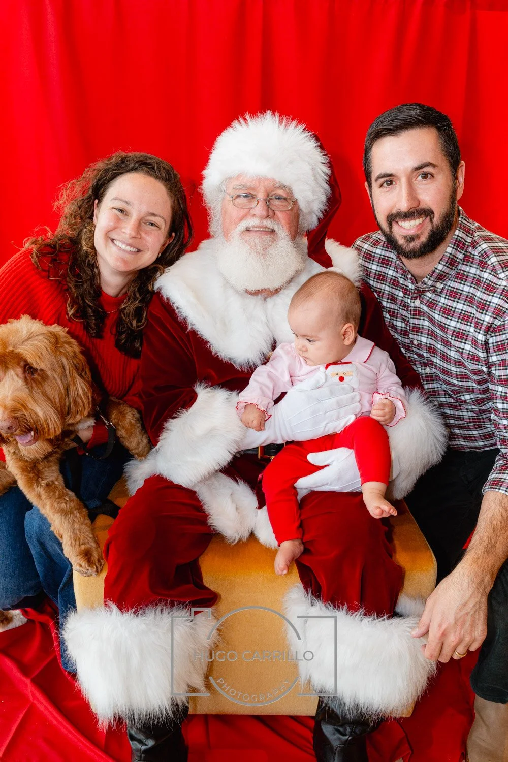 Family gathering with Santa Claus, a young girl, a man, a woman, and a dog in front of a red curtain.