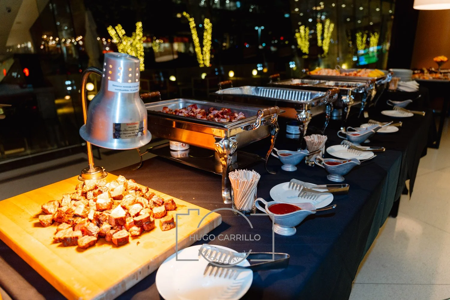 Buffet table with chafing dishes containing cooked meat, small bowls of red sauce, and bowls of toothpicks, set up in a restaurant or event space with city lights visible through the windows in the background.