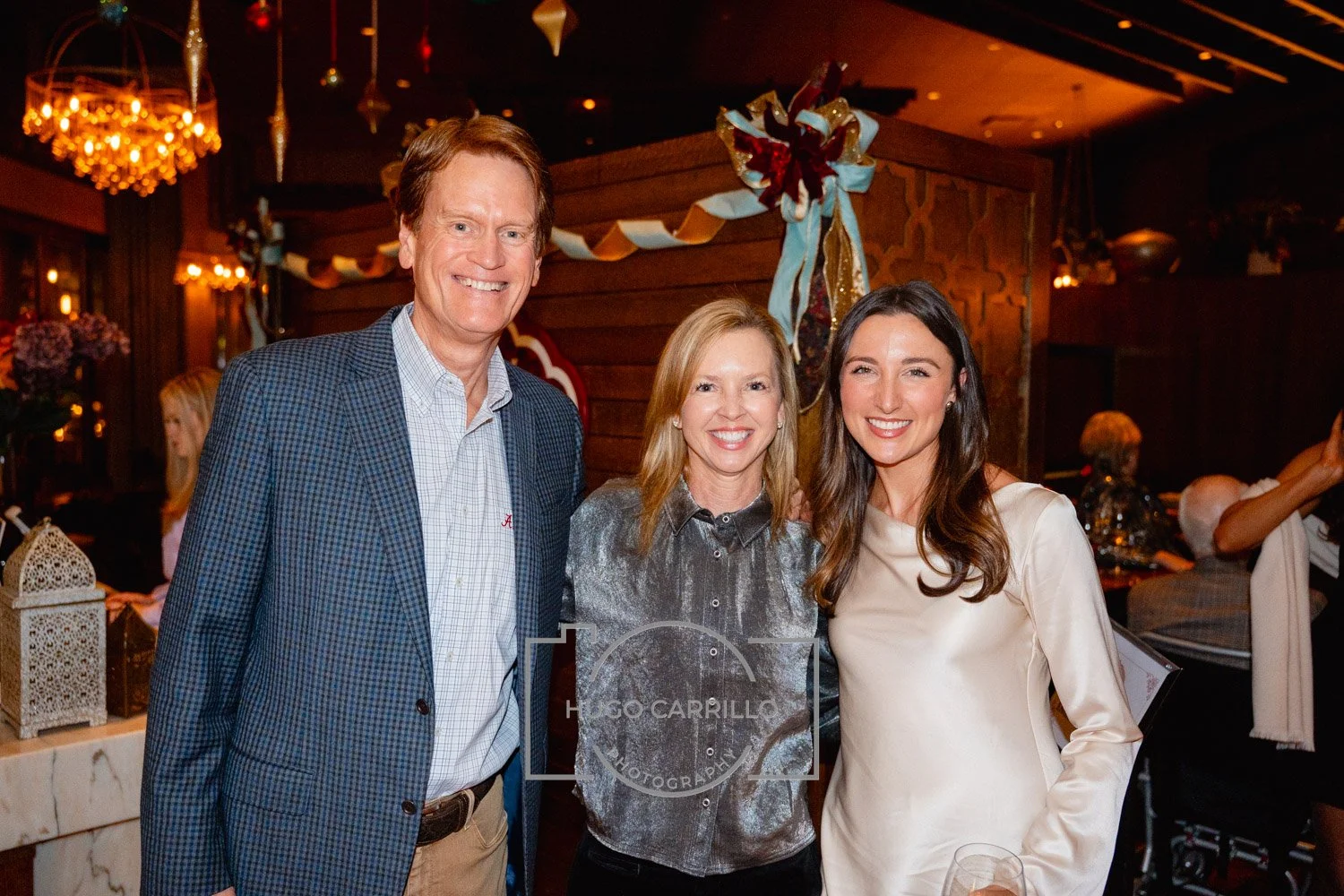 Three people smiling at an indoor event, standing close together in a warmly lit room with holiday decorations in the background.
