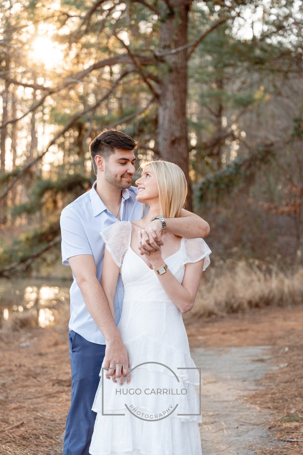 A couple stands close together outdoors in a forested area with sunlight filtering through trees. The woman wears a white dress with lace sleeves, and the man wears a light blue shirt and dark pants. They look into each other's eyes affectionately.