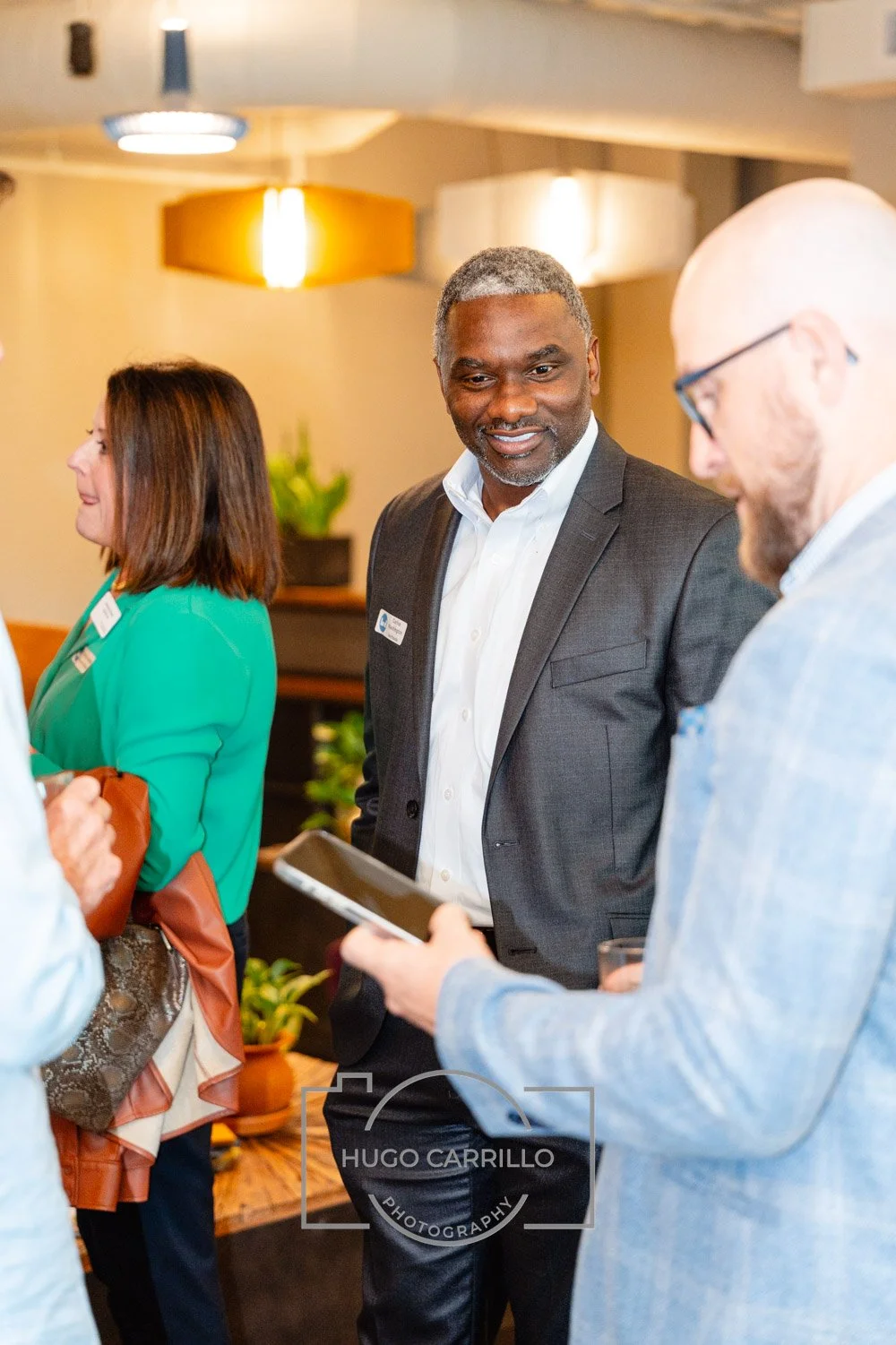 A group of professionals at a networking event, engaging in conversation. The central figure is a middle-aged Black man in a suit, smiling. Others include a woman in a green blazer and a man with glasses and a light-colored blazer. They are indoors w