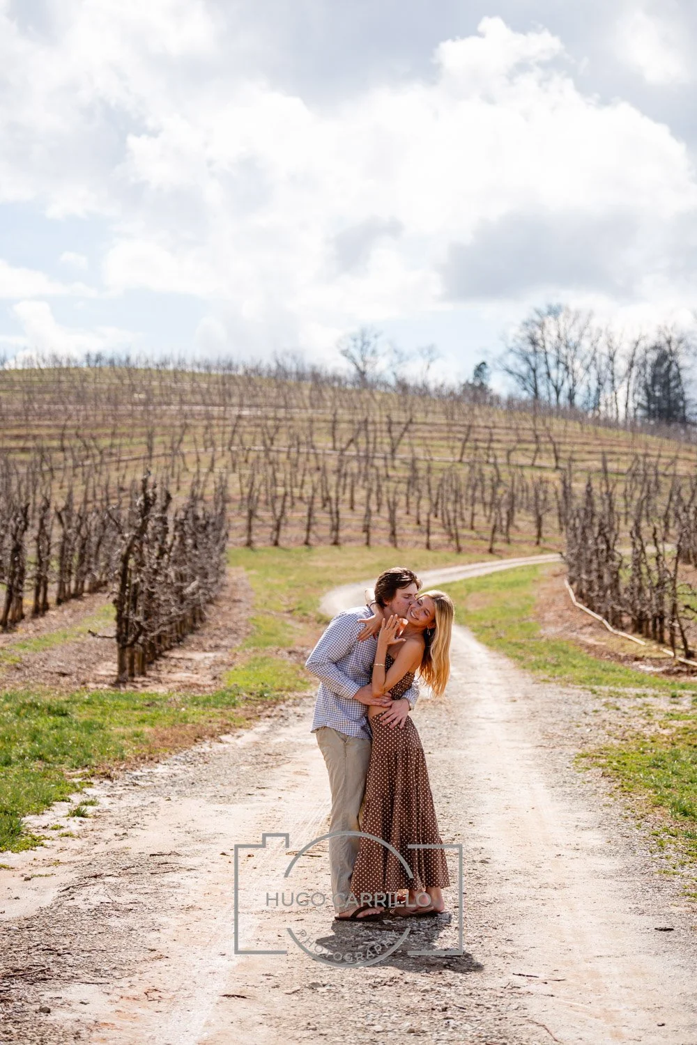 A couple sharing a kiss on a dirt road in a vineyard during daytime, with vines and a cloudy sky in the background.