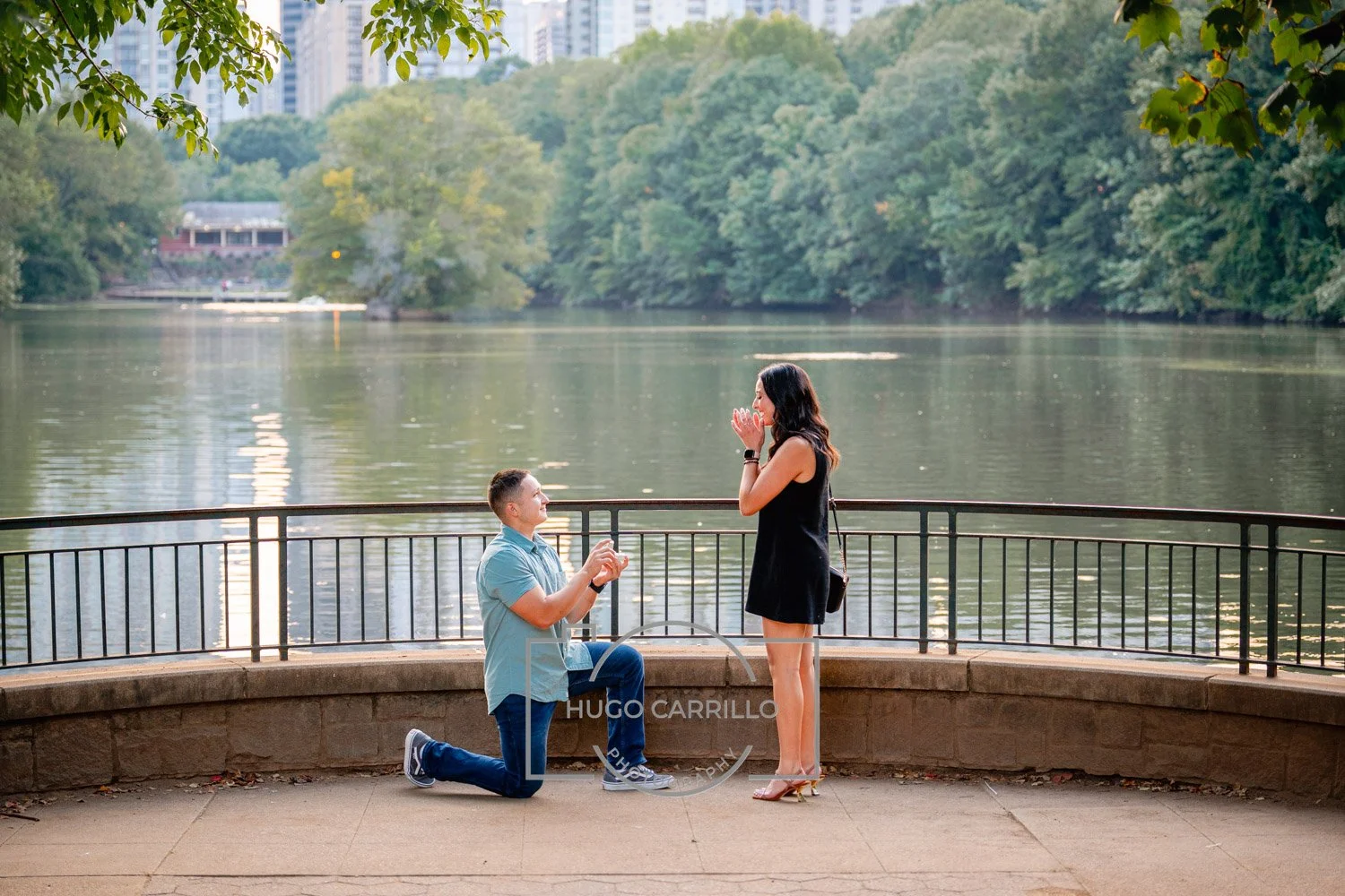 A man kneeling on one knee proposing to a woman at a park by a lake, with trees and city buildings in the background.