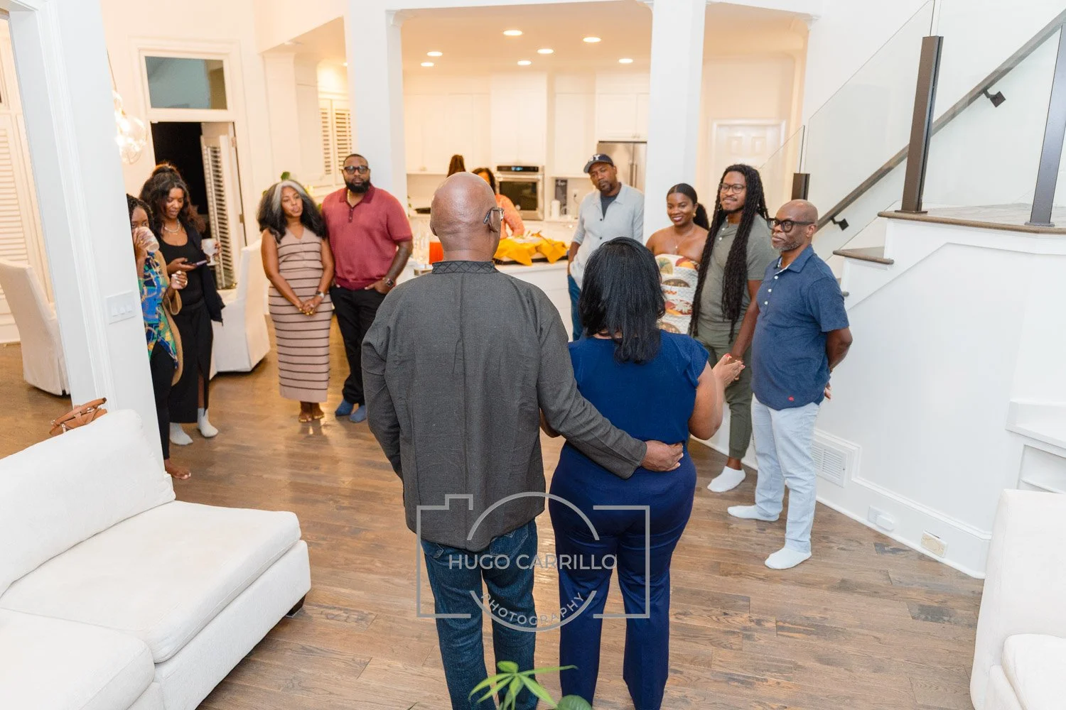 A group of people gathered in a living room with wooden floors, white walls, and an open kitchen in the background. The people are standing in a semi-circle, some smiling and talking, with a couple embracing in the foreground.