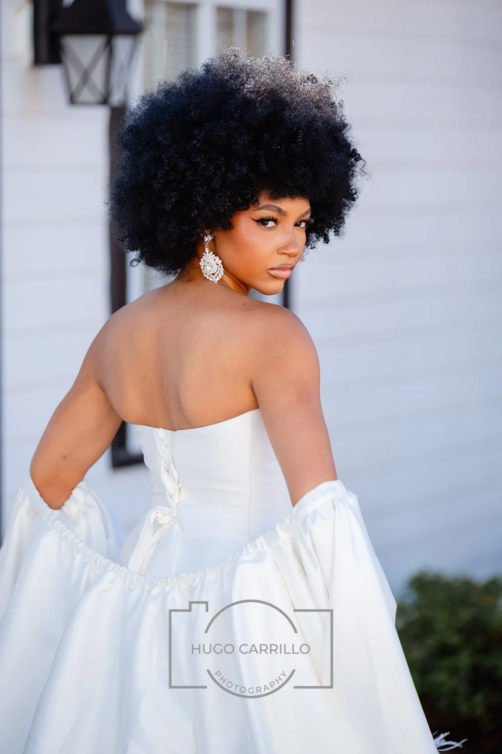 A woman with curly black hair wearing a white strapless dress and large, dangling earrings, posing outdoors by a white building with black window frames.