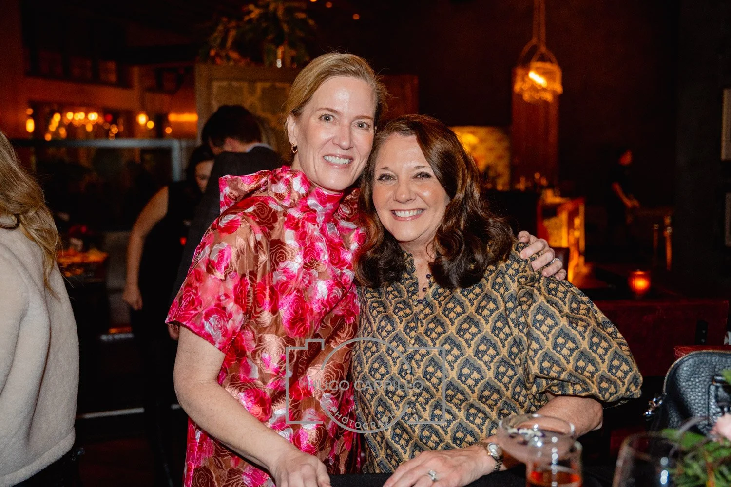 Two women smiling and hugging at a social gathering in a dimly lit venue.