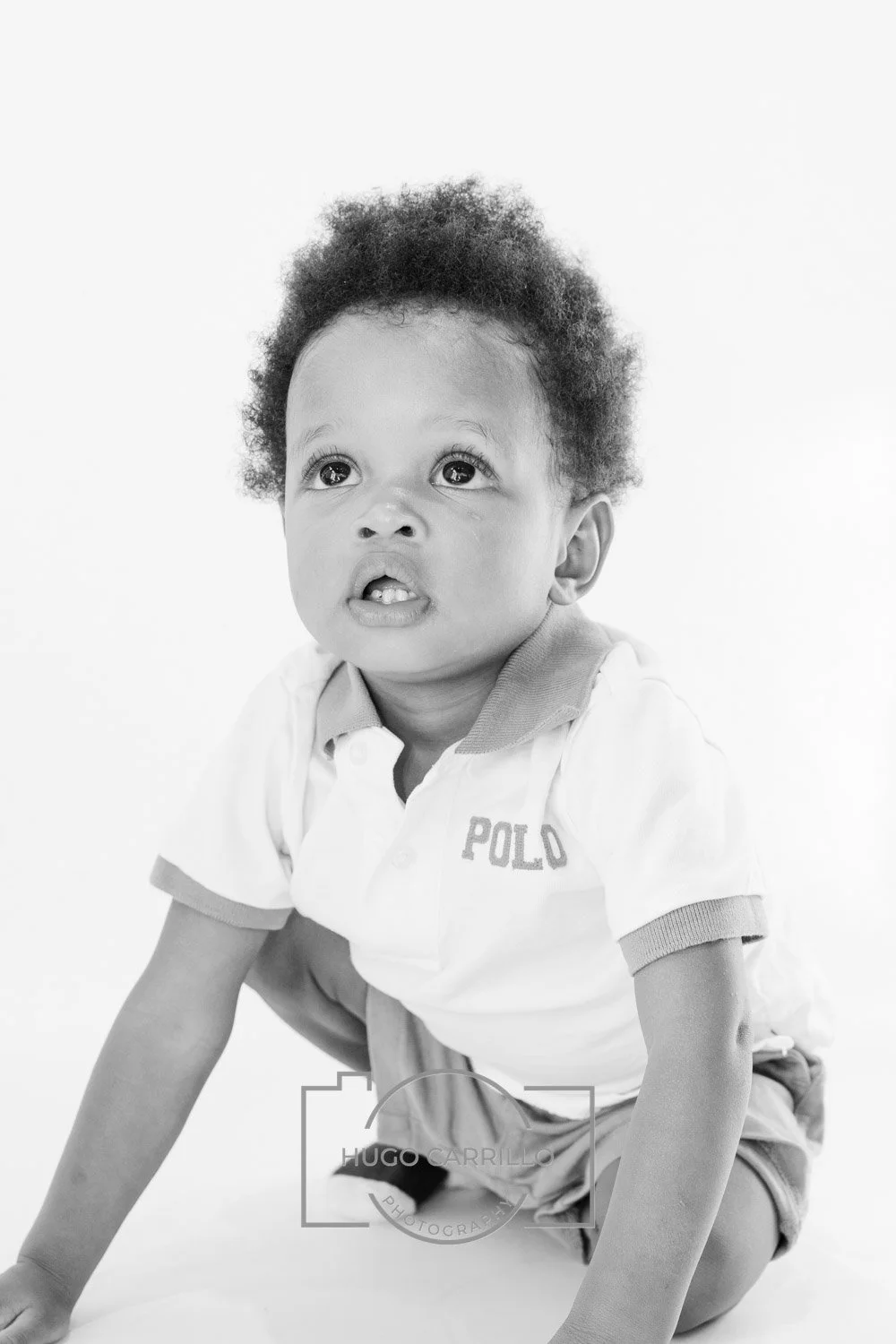 Black and white photo of a young child with curly hair, sitting on the floor and looking up with a curious expression. The child is wearing a polo shirt and shorts.