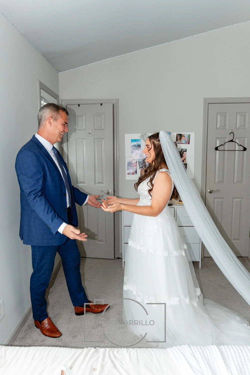 A bride in a white wedding dress and veil happily hands a ring to a man in a blue suit during a wedding or vow exchange in a room with gray walls and closed gray doors.