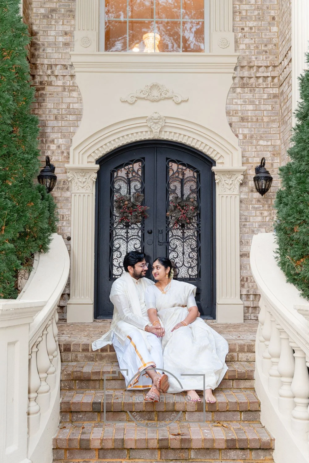 A couple in traditional white Indian attire sitting on steps in front of a decorative black iron door with wreaths, surrounded by white railings and greenery.