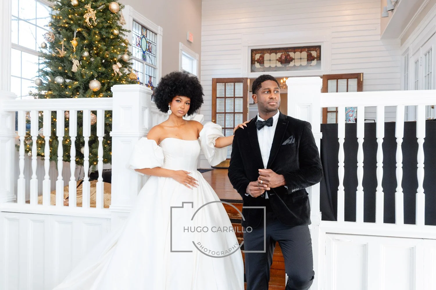 A bride in a white wedding dress with puffed sleeves posing beside a groom in a black tuxedo, inside a decorated venue with a Christmas tree in the background.
