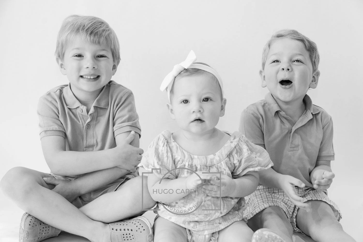 Black and white photo of three children sitting together, two boys on either side of a baby girl, all smiling and engaged in different expressions.