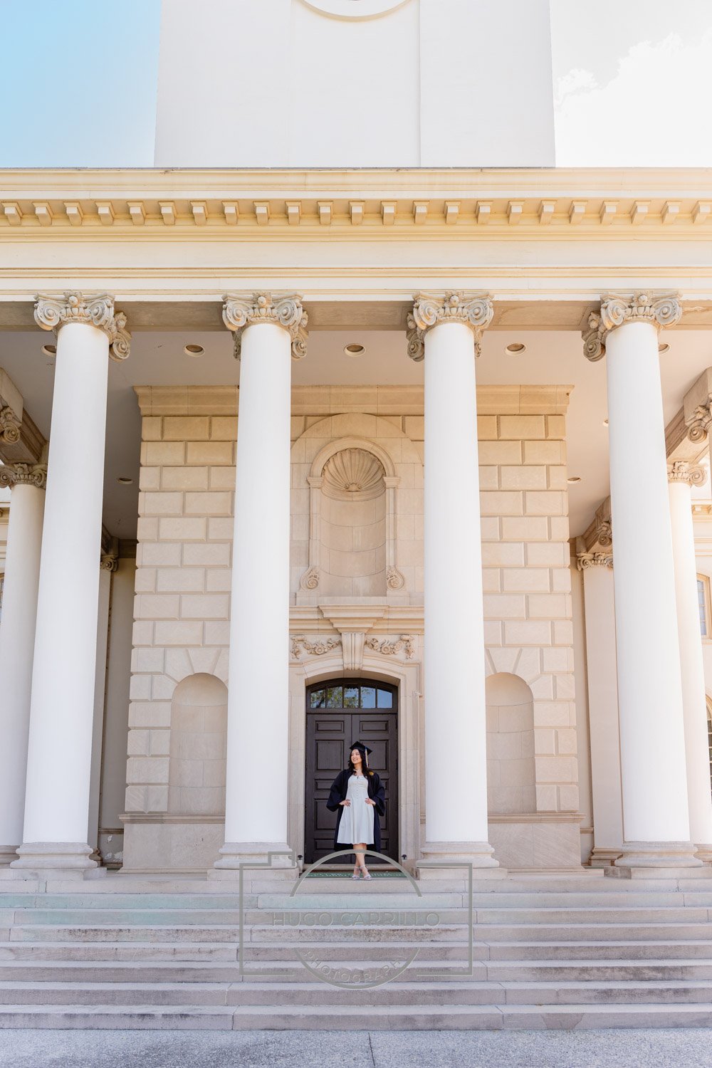 A woman in graduation attire standing on the steps of a grand building with large columns and ornate architecture.