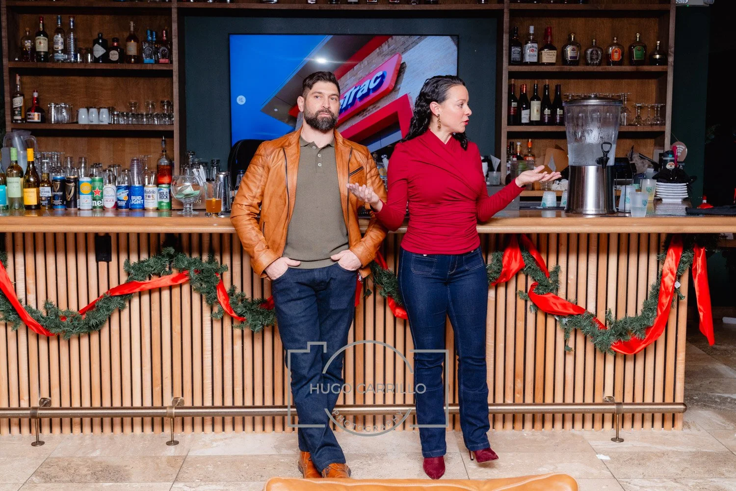 A man and a woman standing at a decorated bar counter, with the woman gesturing with her hands, in a bar or restaurant setting with holiday decorations.