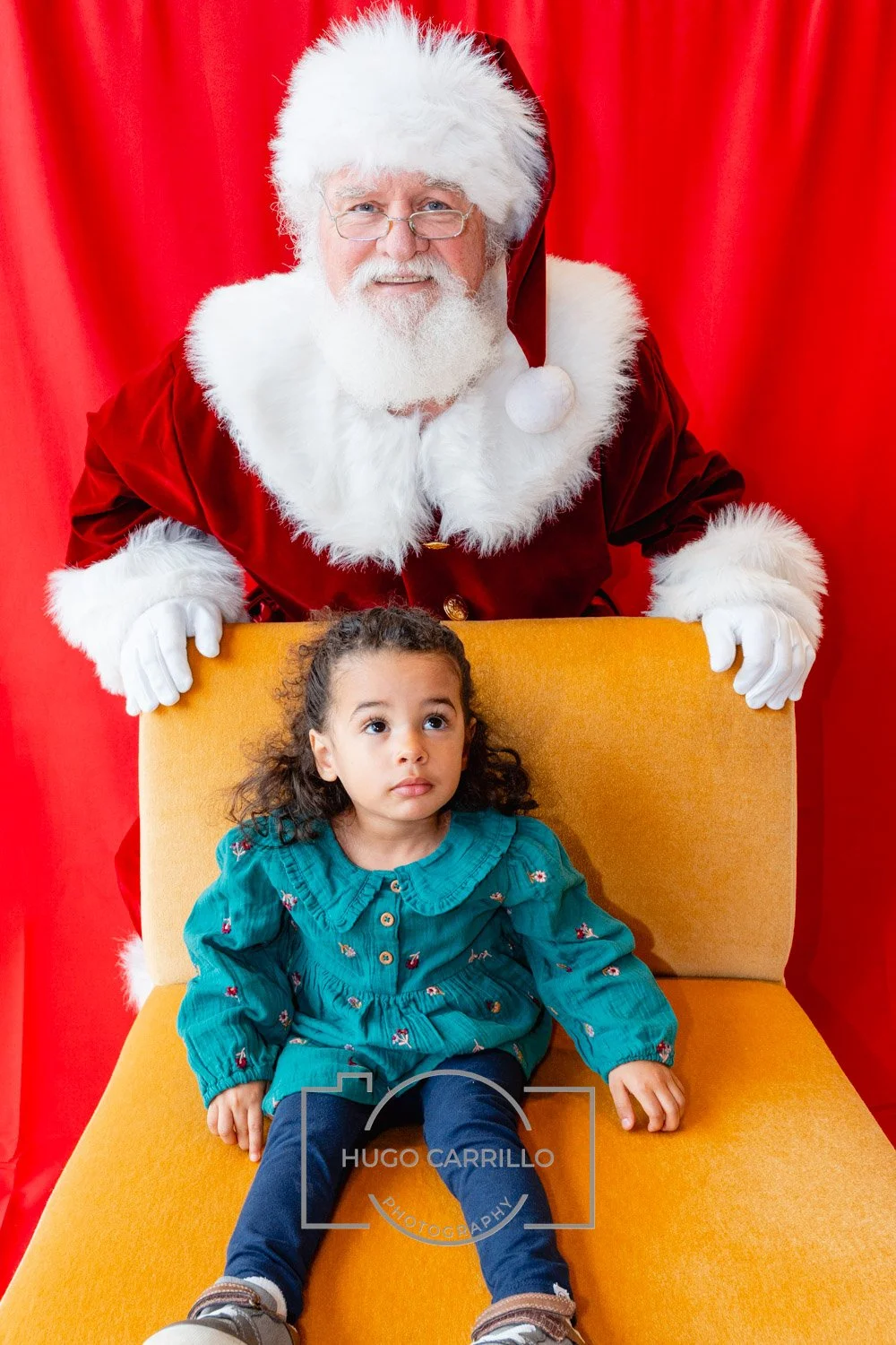 Santa Claus in a red suit with white fur trim, standing behind a yellow chair with a young girl sitting on it, against a red curtain backdrop.