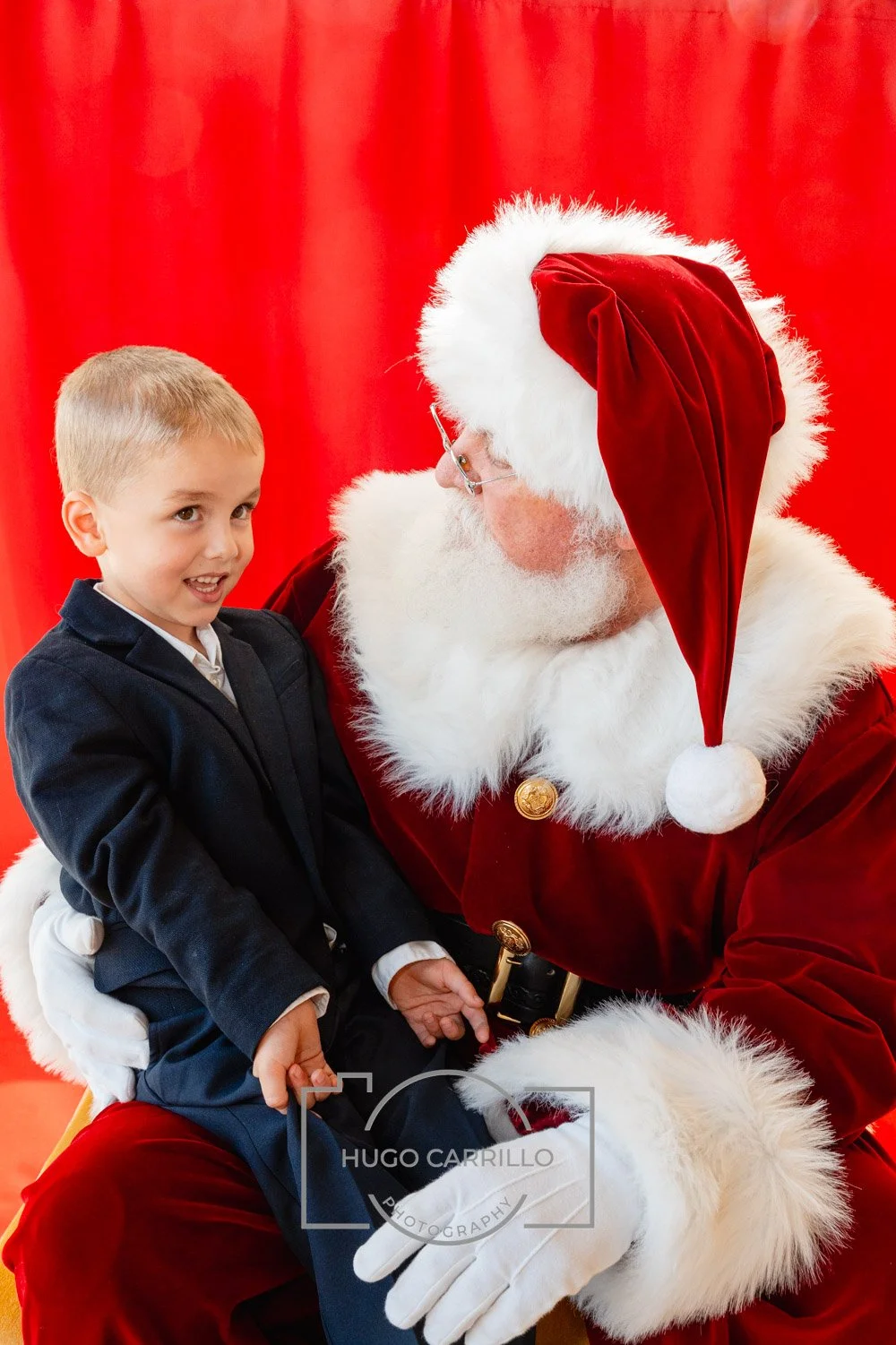 A young boy sitting on Santa Claus's lap, smiling and looking at the camera. Santa is dressed in a traditional red suit with a white fur trim, wearing glasses, and looking at the boy. The background is a red curtain.