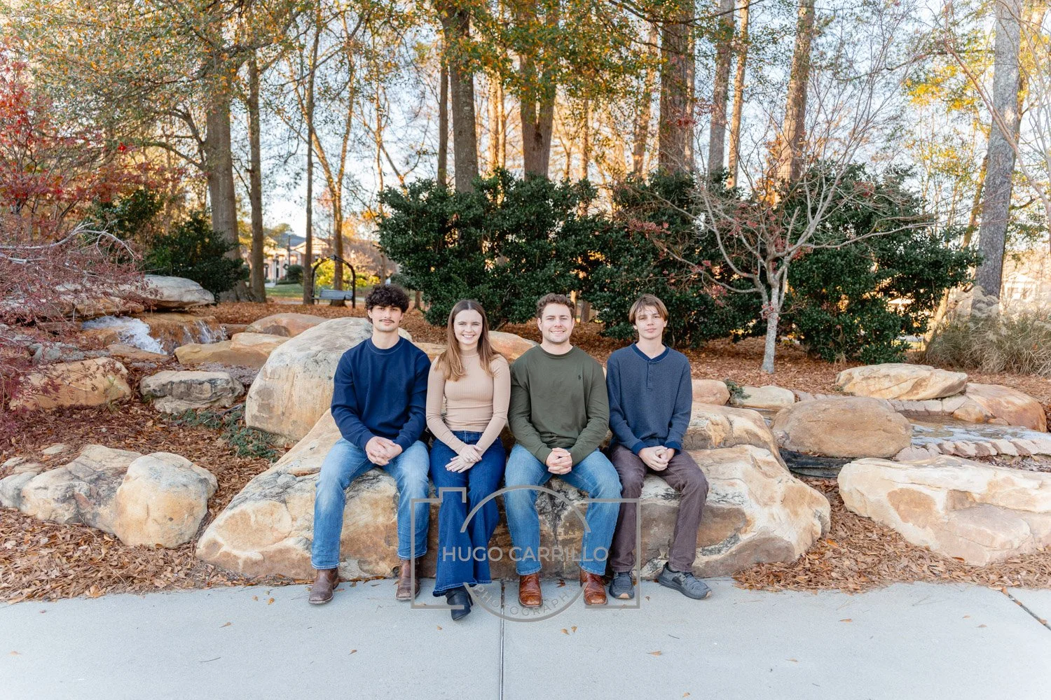 Four young adults sitting on large rocks in a park with trees and autumn foliage in the background during late afternoon.