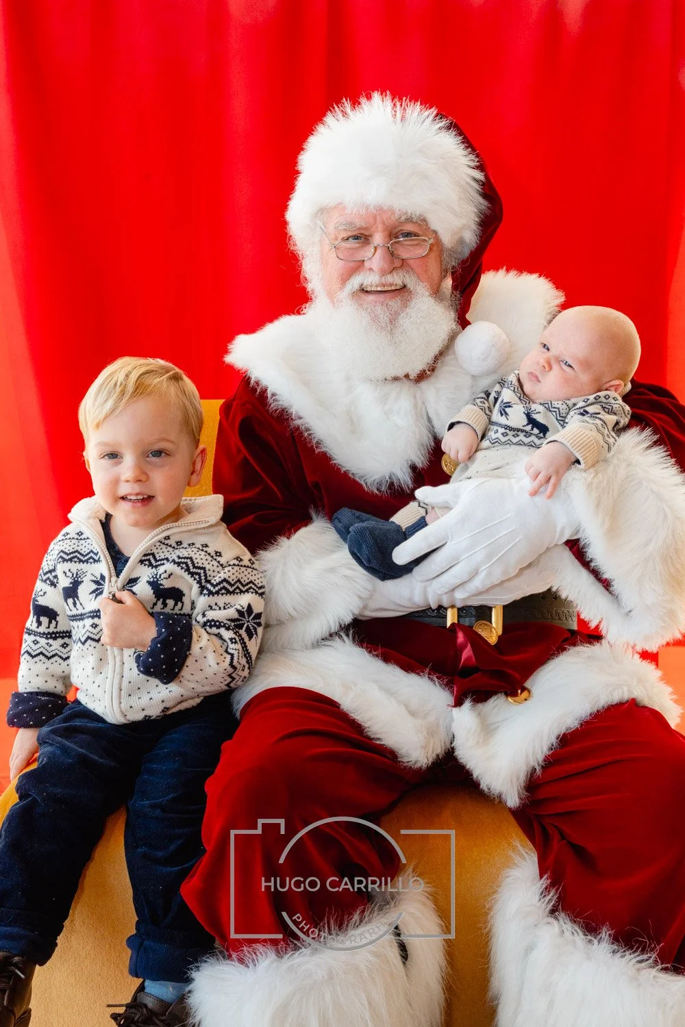 Santa Claus dressed in traditional red and white, holding a baby with a knit sweater, sitting beside a young boy wearing a Christmas-themed sweater, in front of red curtains.