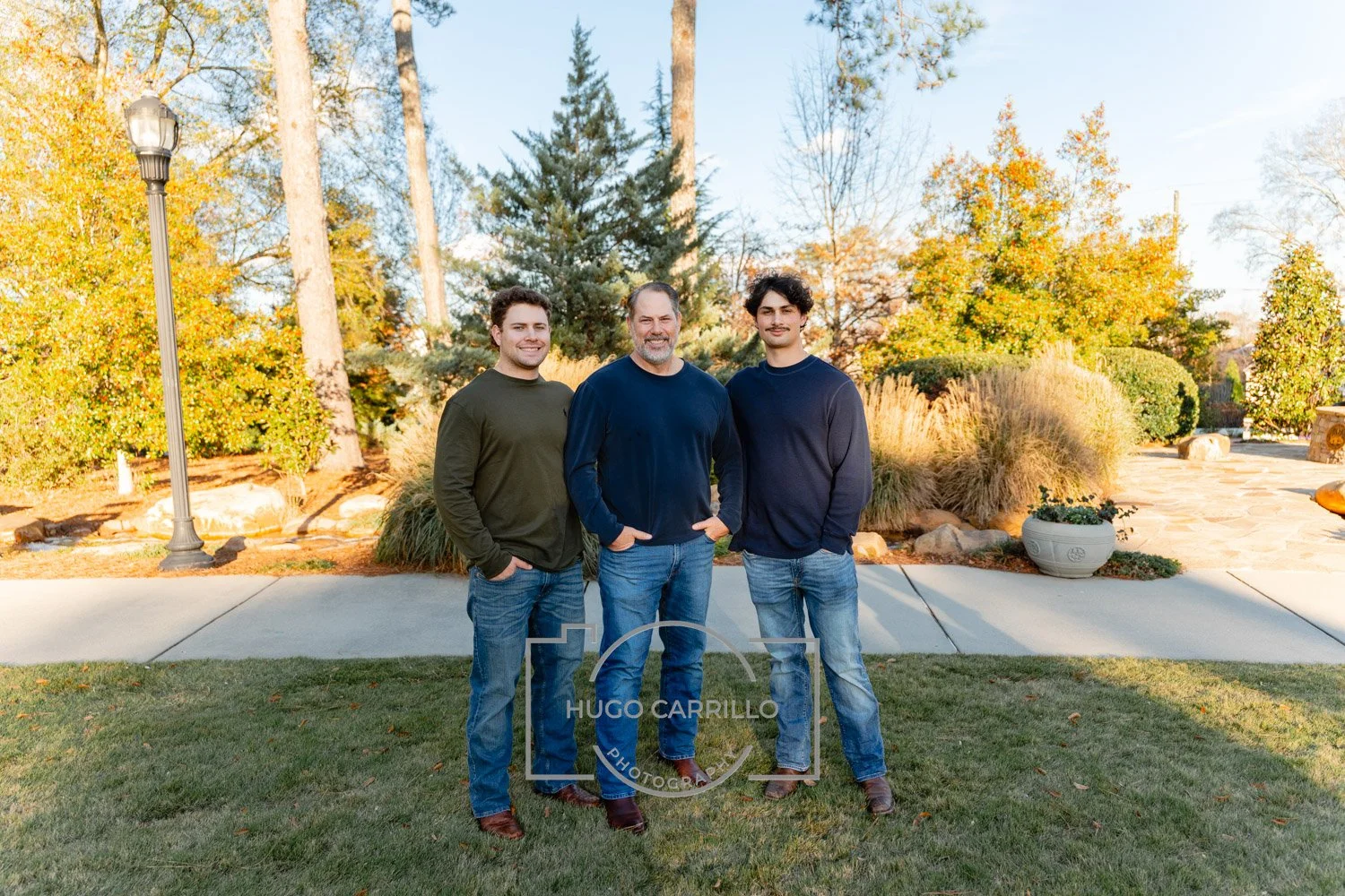 Three men standing outdoors on a grassy area with trees and shrubs in the background. They are posing for a photo, smiling, and wearing casual clothing.