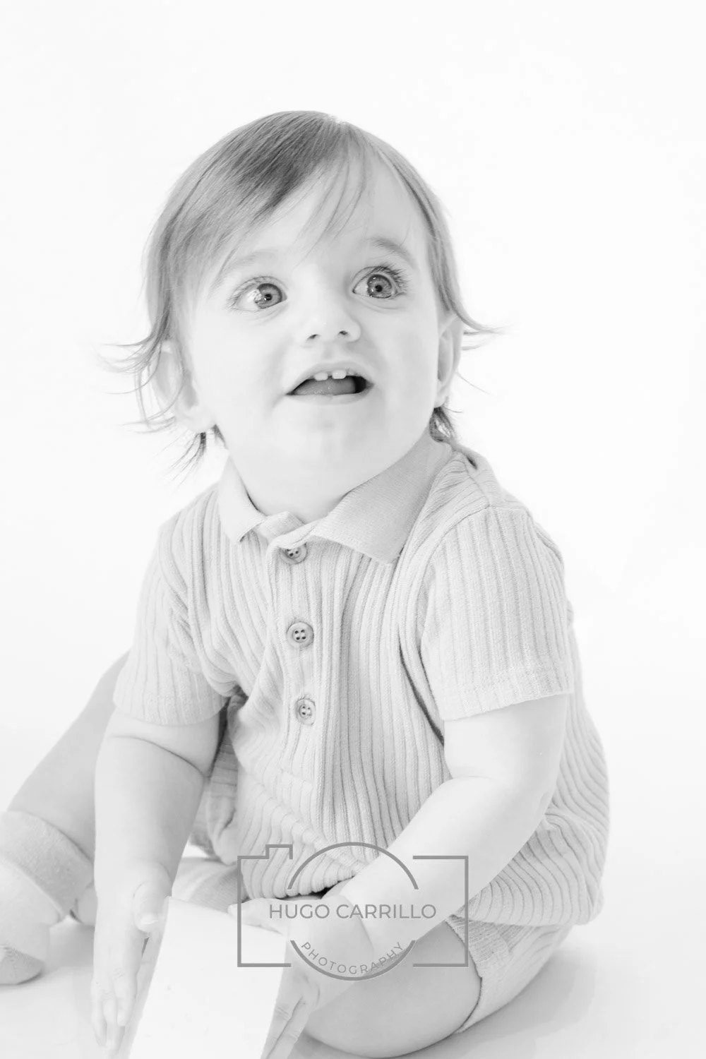 Black and white photo of a toddler with wide eyes and an open mouth, wearing a striped button-up shirt and shorts, sitting on the floor against a plain background.